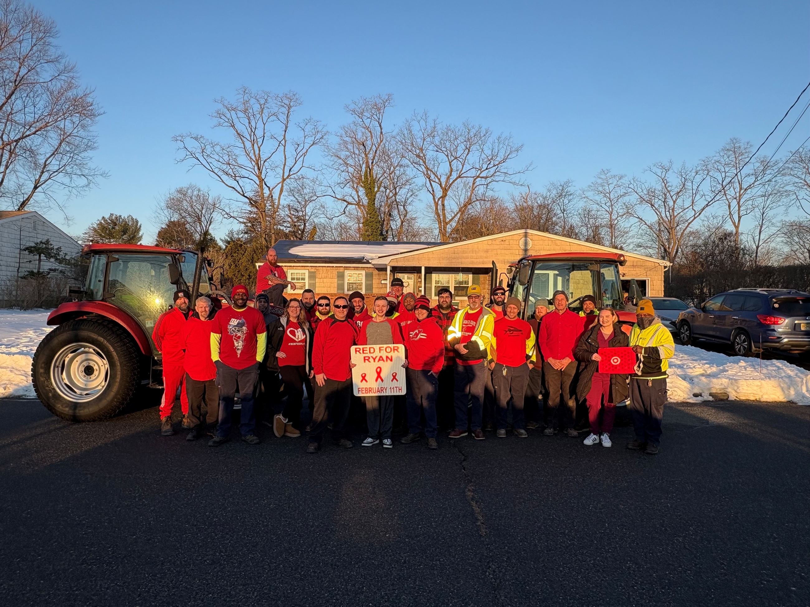 Group in red shirts poses with two tractors outdoors; banner says “RED FOR RYAN” and “FEBRUARY 11TH.”